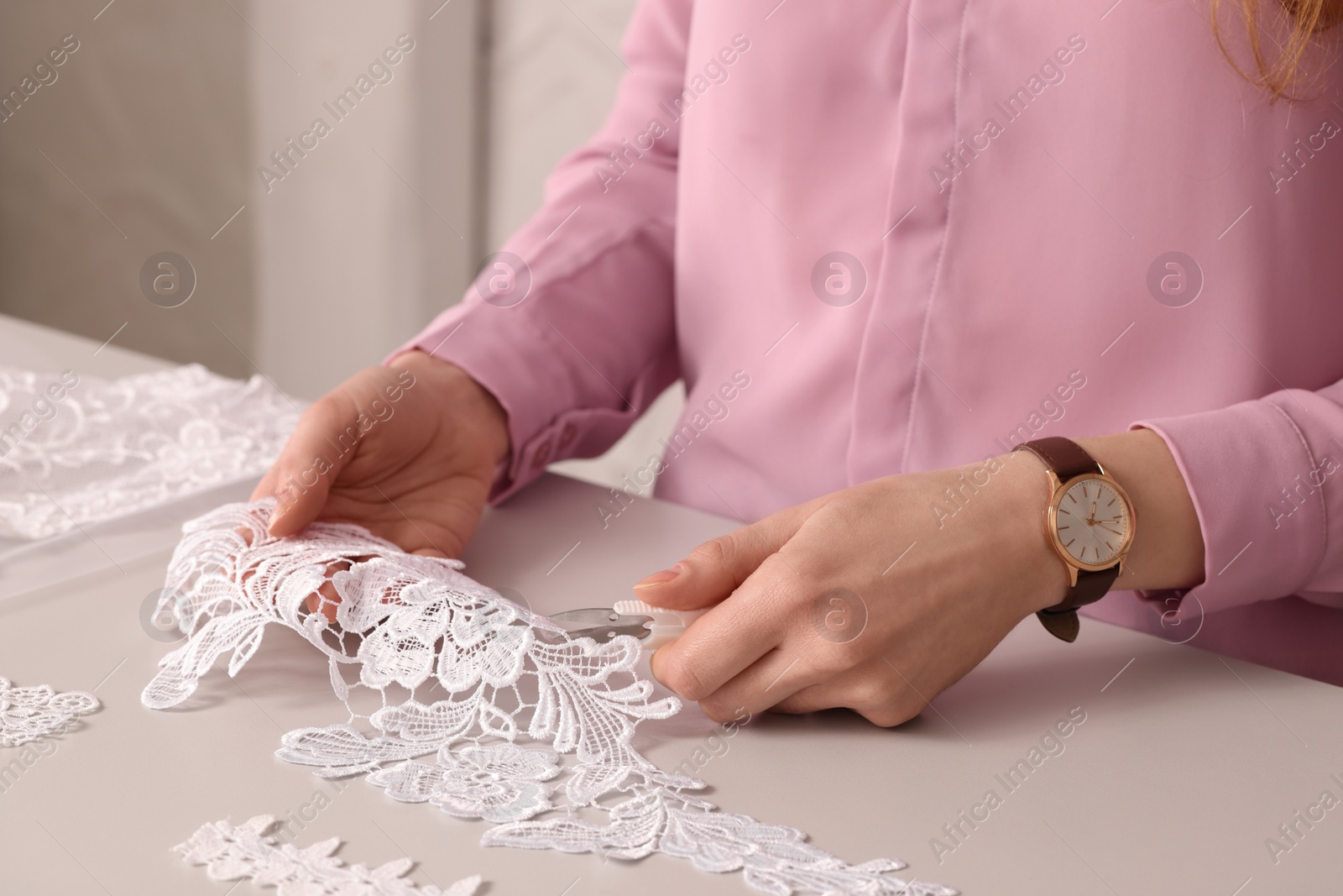 Photo of Dressmaker cutting beautiful white lace at table in atelier, closeup