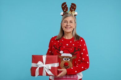 Senior woman in Christmas sweater and reindeer headband holding gift on light blue background