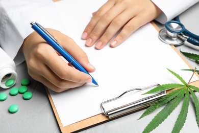 Photo of Female doctor with clipboard, pills and hemp leaf at grey table, closeup