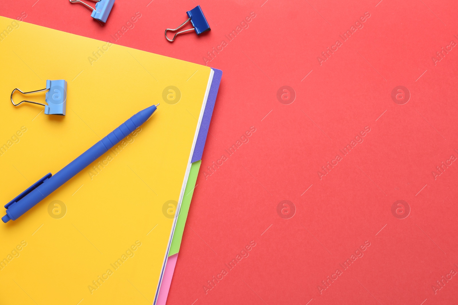 Photo of Yellow notebook with pen and binder clips on red background, flat lay. Space for text