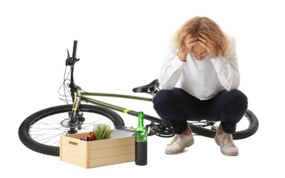 Upset young man with bottle of wine and box of belongings near bicycle on white background