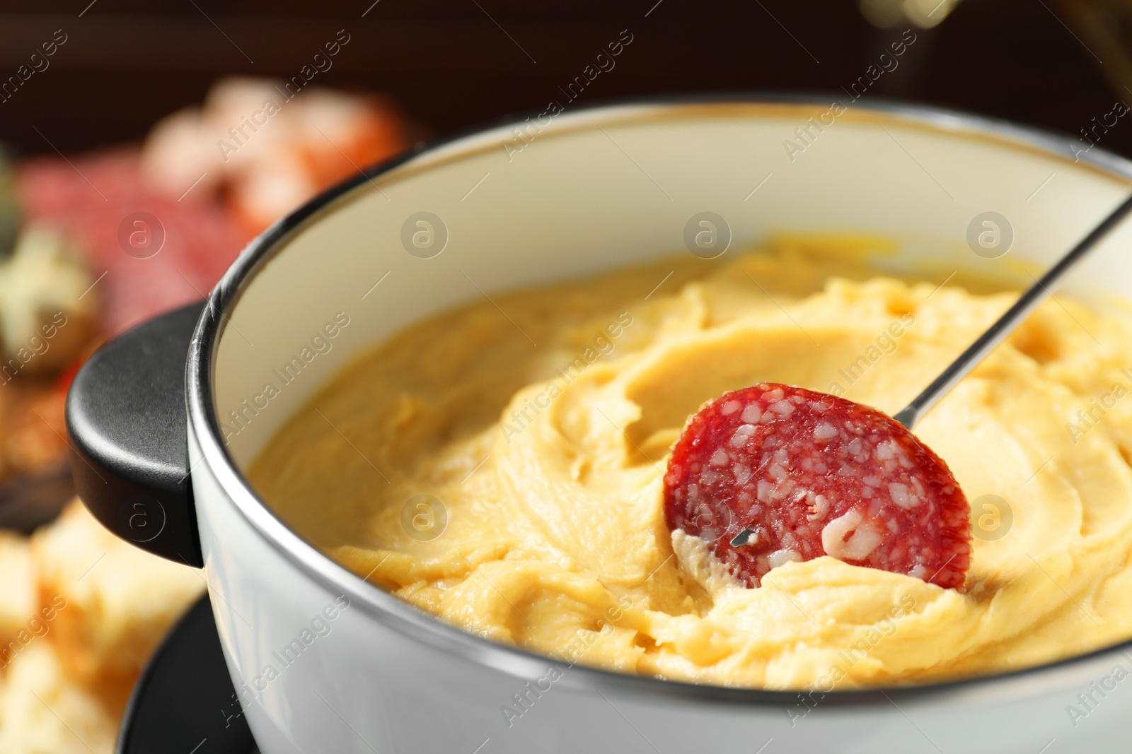 Photo of Dipping piece of sausage into fondue pot with melted cheese on blurred background, closeup