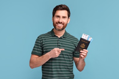 Smiling man pointing at passport and tickets on light blue background