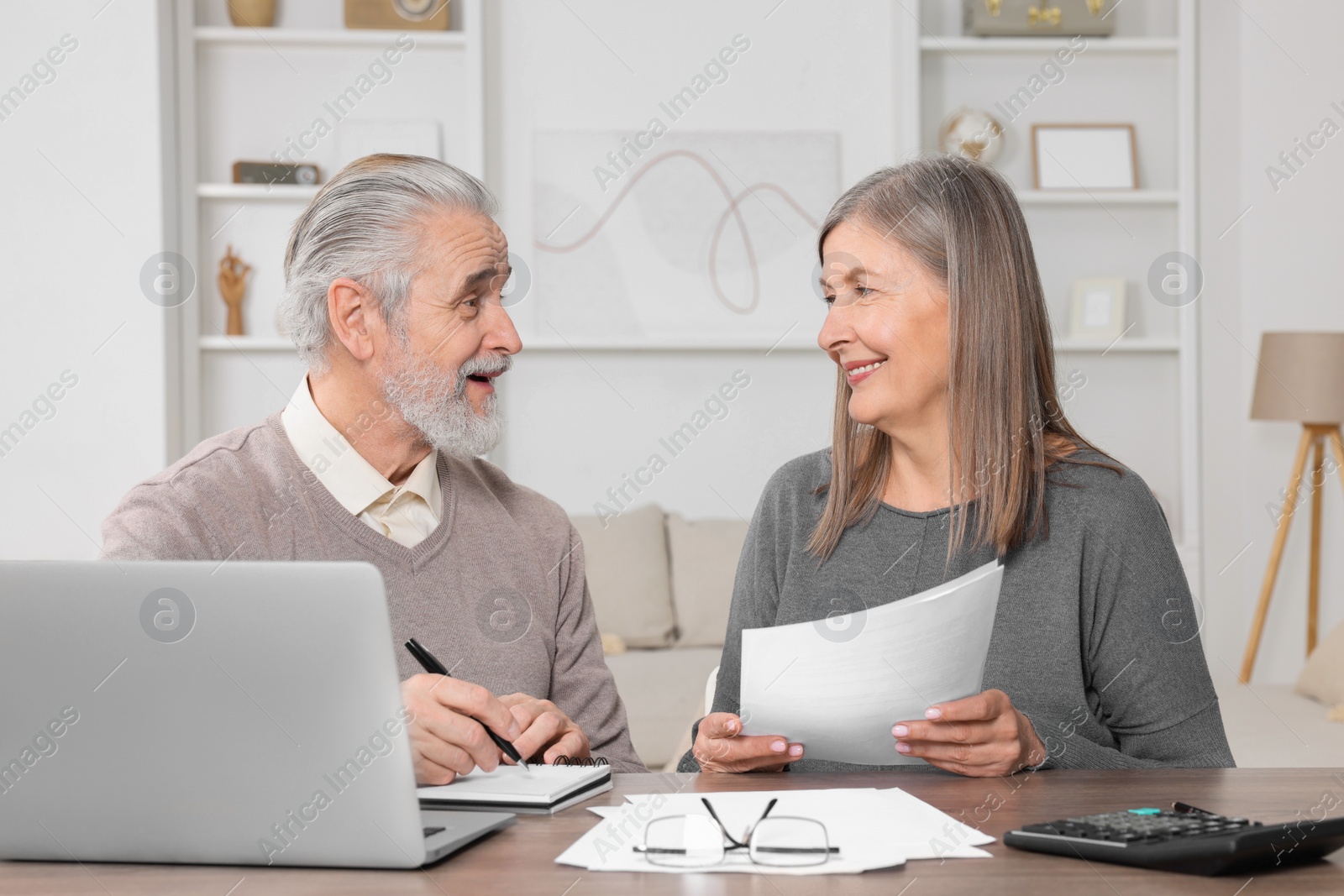 Photo of Elderly couple with papers and laptop discussing pension plan at wooden table in room