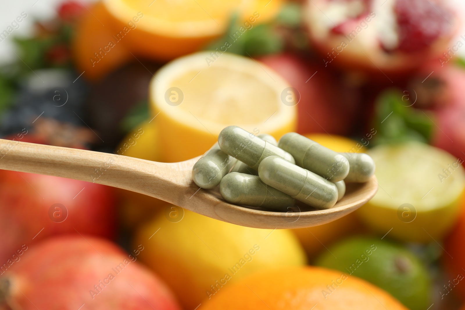 Photo of Vitamin pills in spoon against fresh fruits, closeup