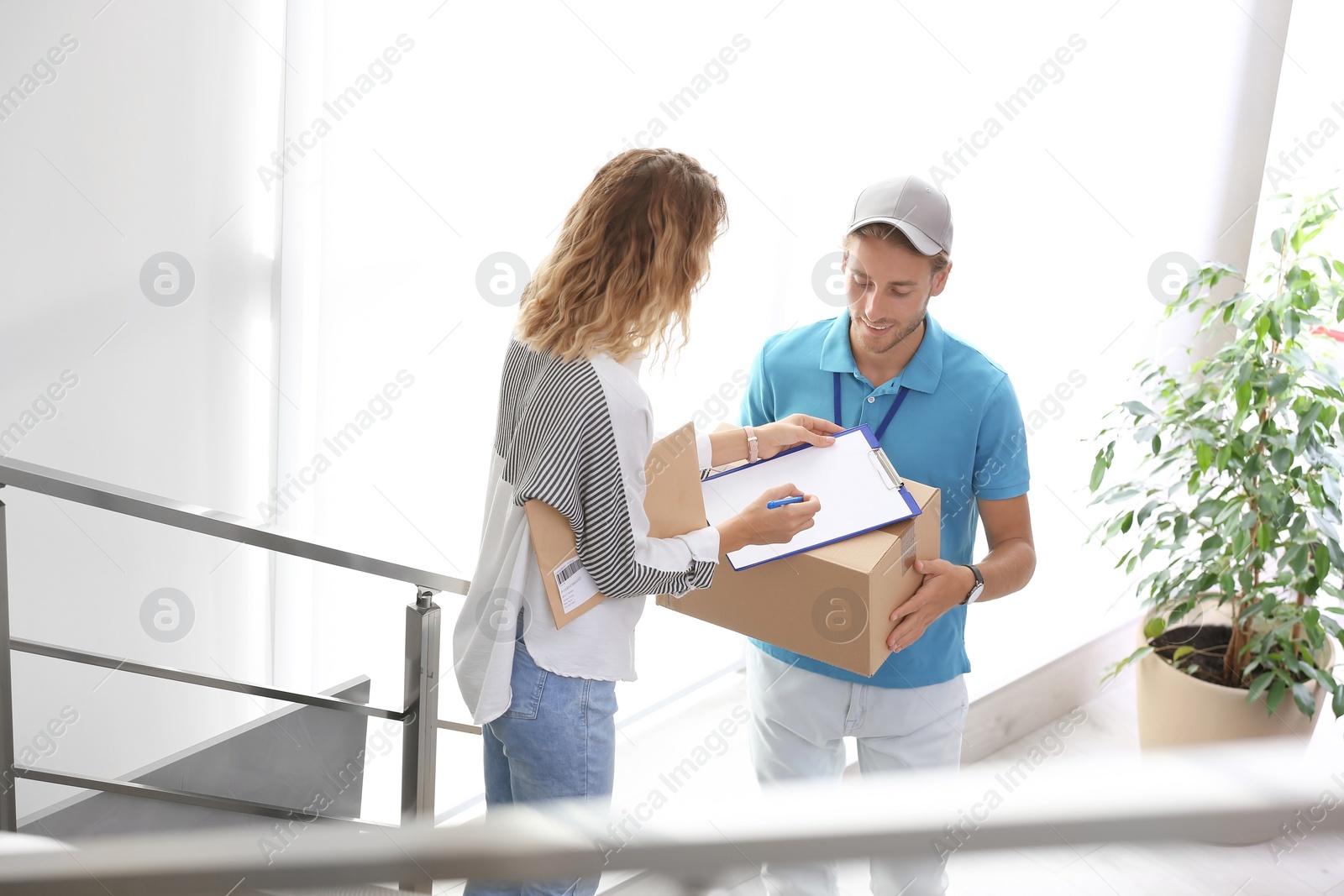 Photo of Young woman signing documents after receiving parcels from courier indoors