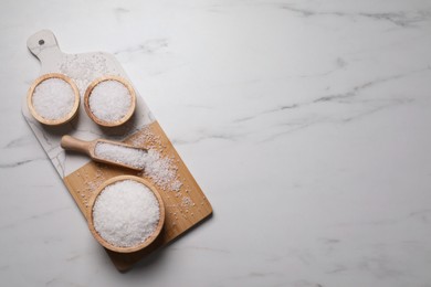 Photo of Wooden board with natural sea salt in scoop and bowls on white marble table, flat lay. Space for text