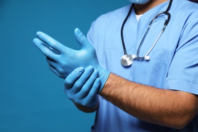 Doctor putting on medical gloves against blue background, closeup
