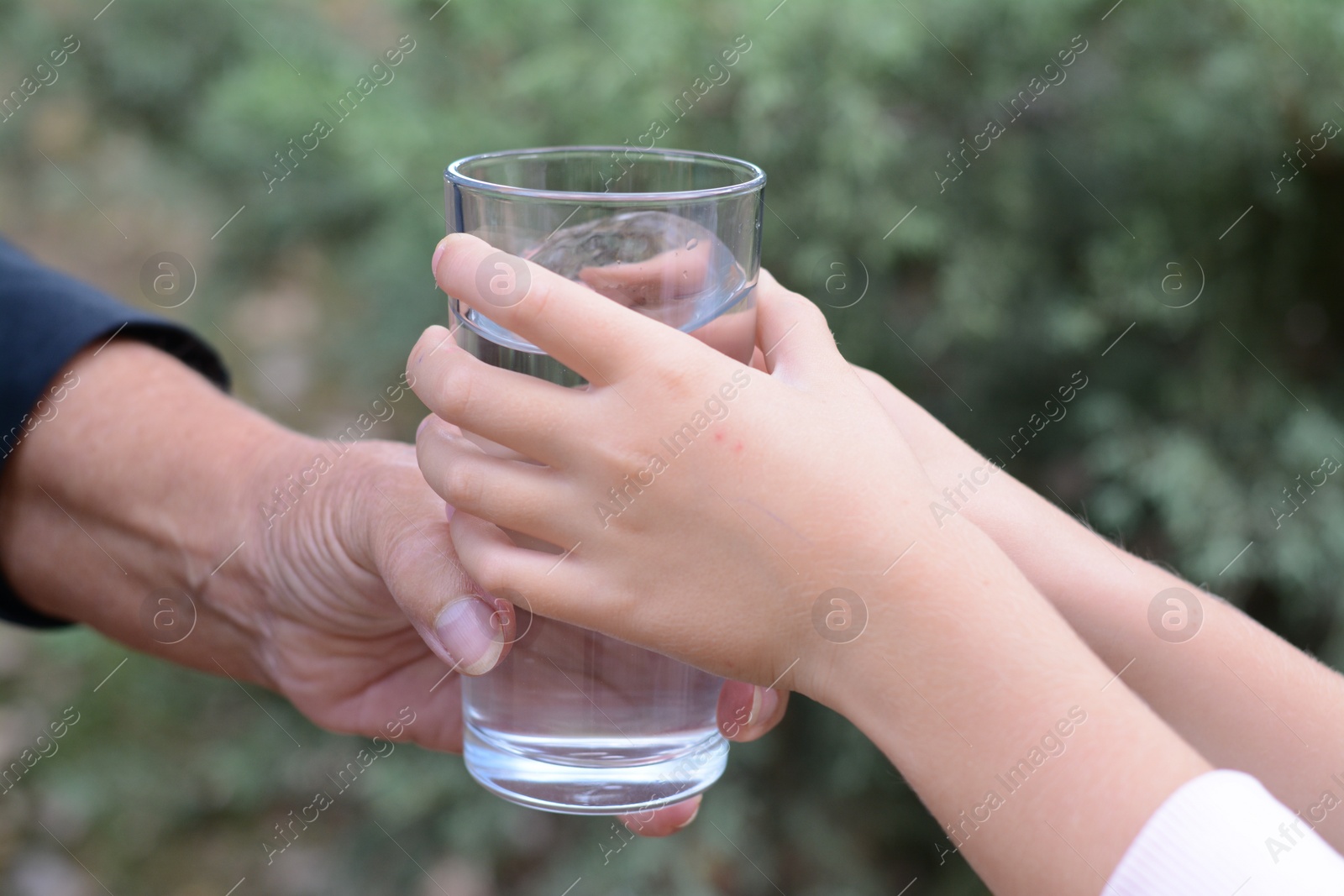 Photo of Child giving glass of water to elderly woman outdoors, closeup