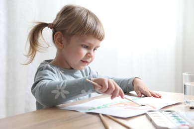 Photo of Cute little girl drawing with brush at wooden table indoors. Child`s art