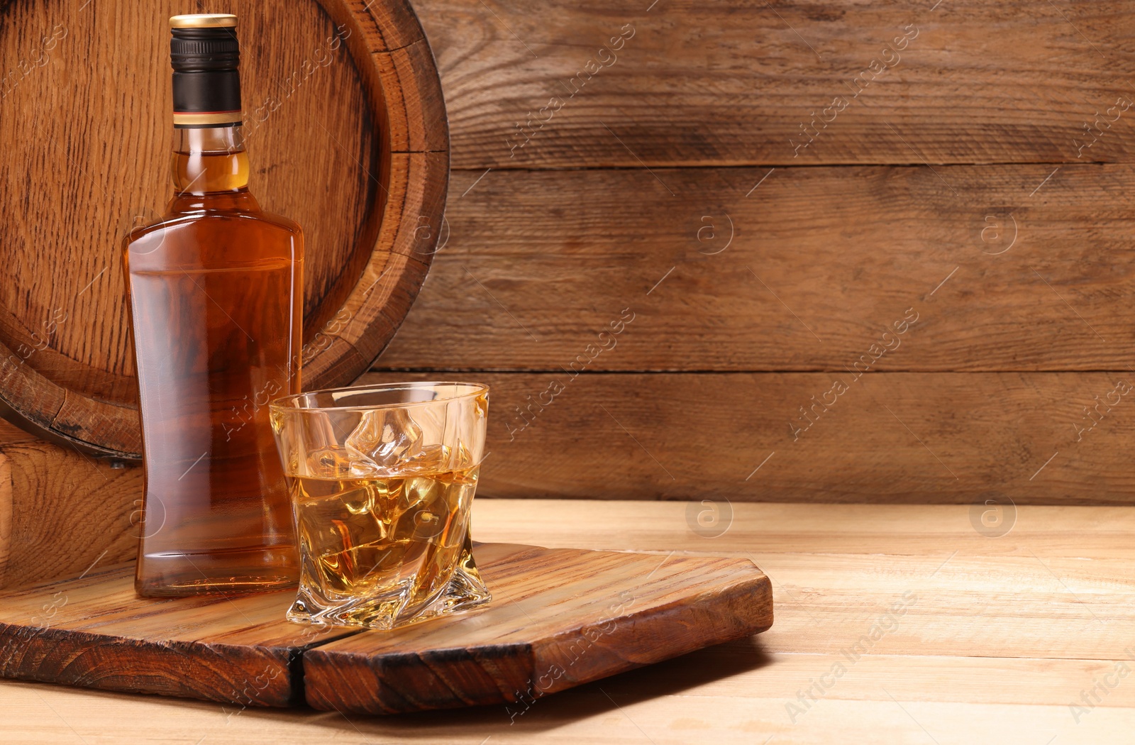 Photo of Whiskey with ice cubes in glass, bottle and barrel on wooden table, space for text