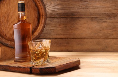 Whiskey with ice cubes in glass, bottle and barrel on wooden table, space for text