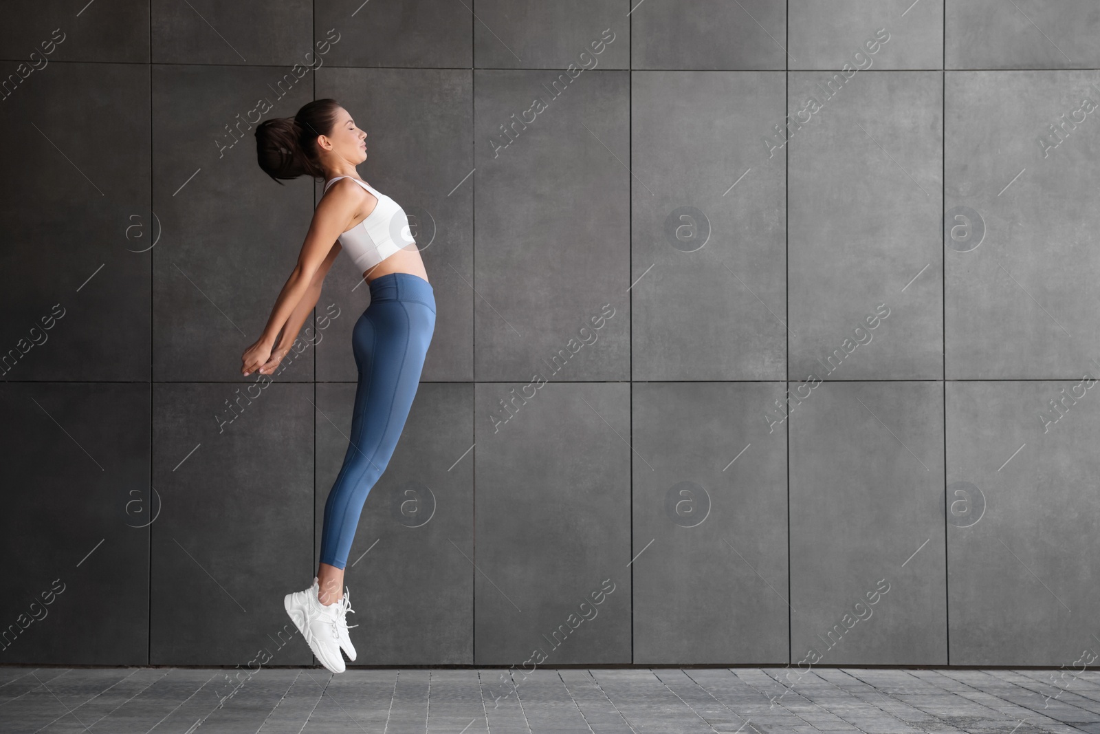 Photo of Beautiful woman in sportswear jumping near dark grey wall outdoors. Space for text