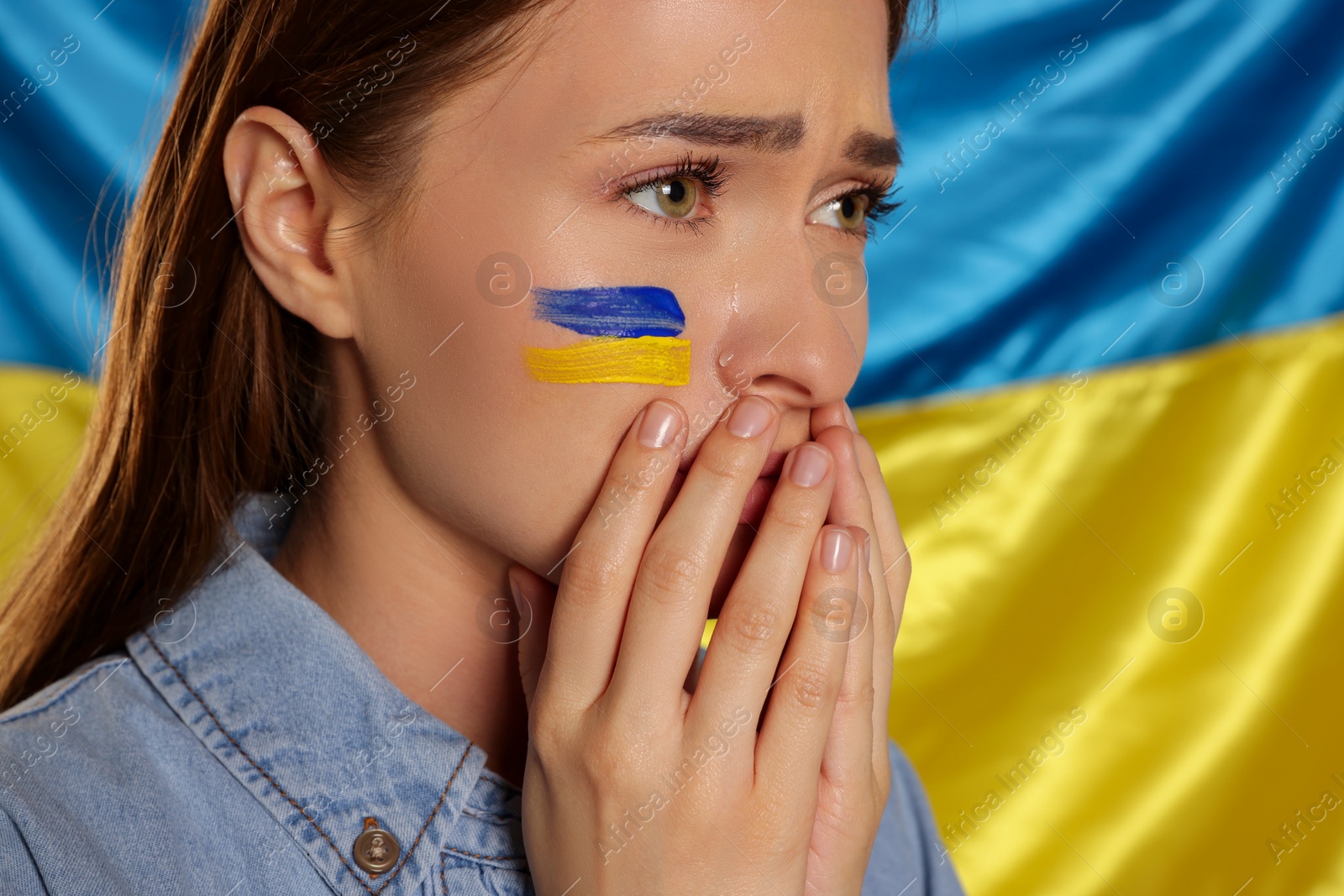 Photo of Sad young woman with clasped hands near Ukrainian flag, closeup. Space for text