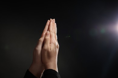 Photo of Woman holding hands clasped while praying against light in darkness, closeup. Space for text