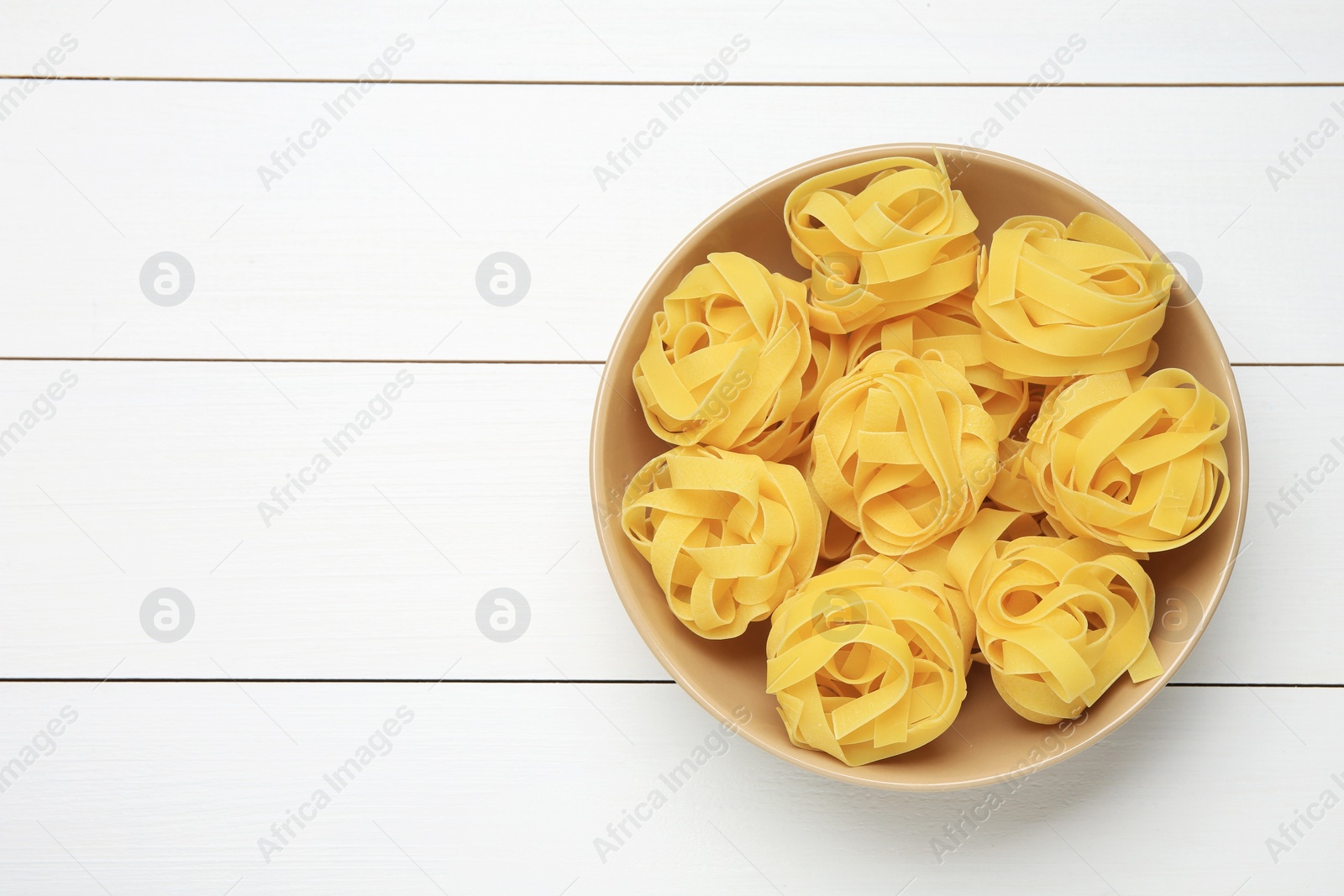 Photo of Raw tagliatelle pasta in bowl on white wooden table, top view. Space for text