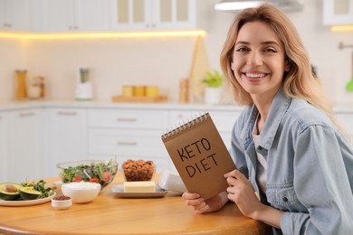 Woman holding notebook with phrase Keto Diet and different products at wooden table in kitchen, space for text