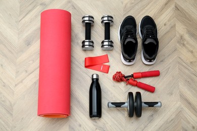 Photo of Exercise mat, dumbbells, bottle of water, skipping rope, ab roller, fitness elastic band and shoes on wooden floor, flat lay