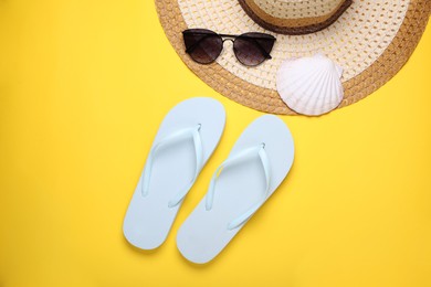 Stylish white flip flops, sunglasses, hat and seashell on yellow background, flat lay