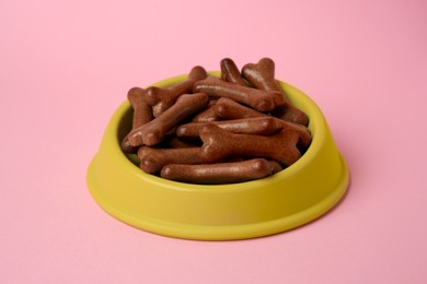 Photo of Yellow bowl with bone shaped dog cookies on pink background, closeup