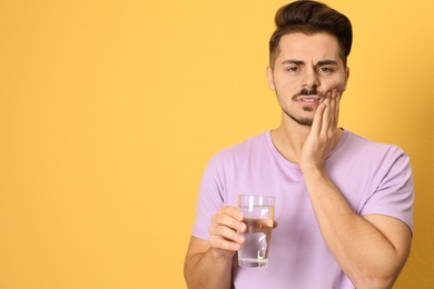 Emotional young man with sensitive teeth and glass of water on color background. Space for text