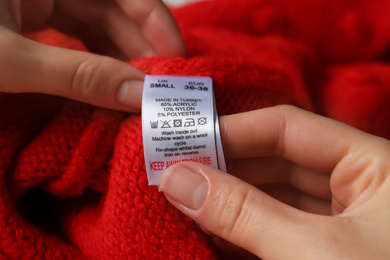 Photo of Woman reading clothing label with care symbols and material content on red knitted sweater, closeup