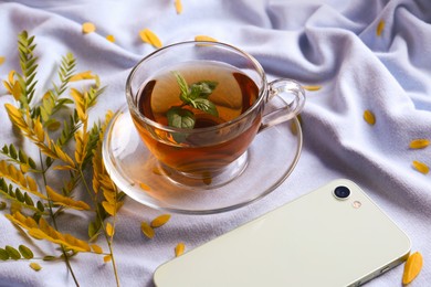 Photo of Cup of aromatic herb tea, smartphone and dry autumn leaves on white cloth