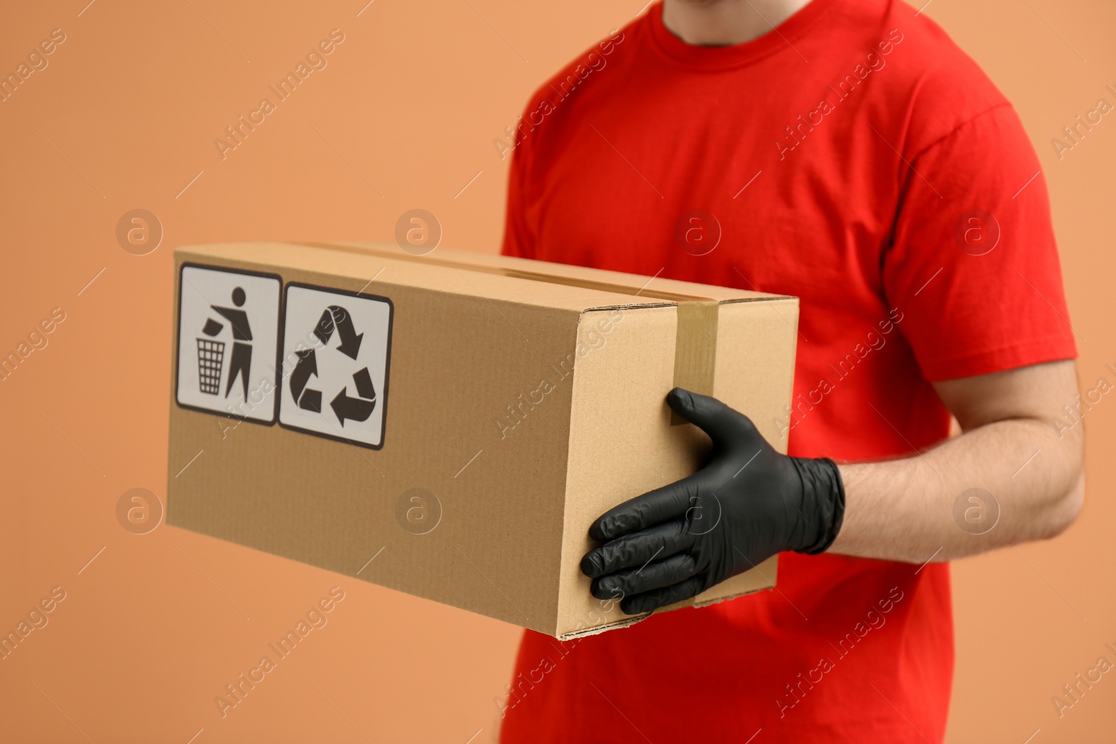 Photo of Courier holding cardboard box with different packaging symbols on orange background, closeup. Parcel delivery