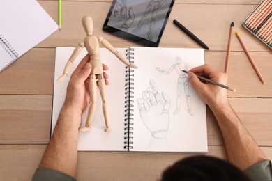 Photo of Man drawing mannequin in sketchbook with pencil at wooden table, top view