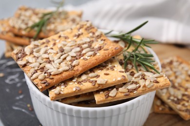 Photo of Cereal crackers with flax, sunflower, sesame seeds and rosemary in bowl on board, closeup
