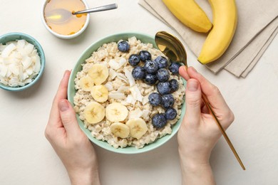Woman eating tasty oatmeal with banana, blueberries, honey and coconut flakes at beige table, top view