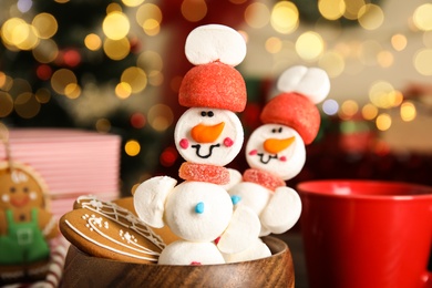 Photo of Funny snowmen made of marshmallows in wooden bowl against blurred festive lights, closeup