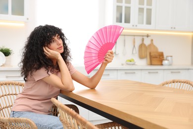 Photo of Young woman waving pink hand fan to cool herself at table in kitchen. Space for text