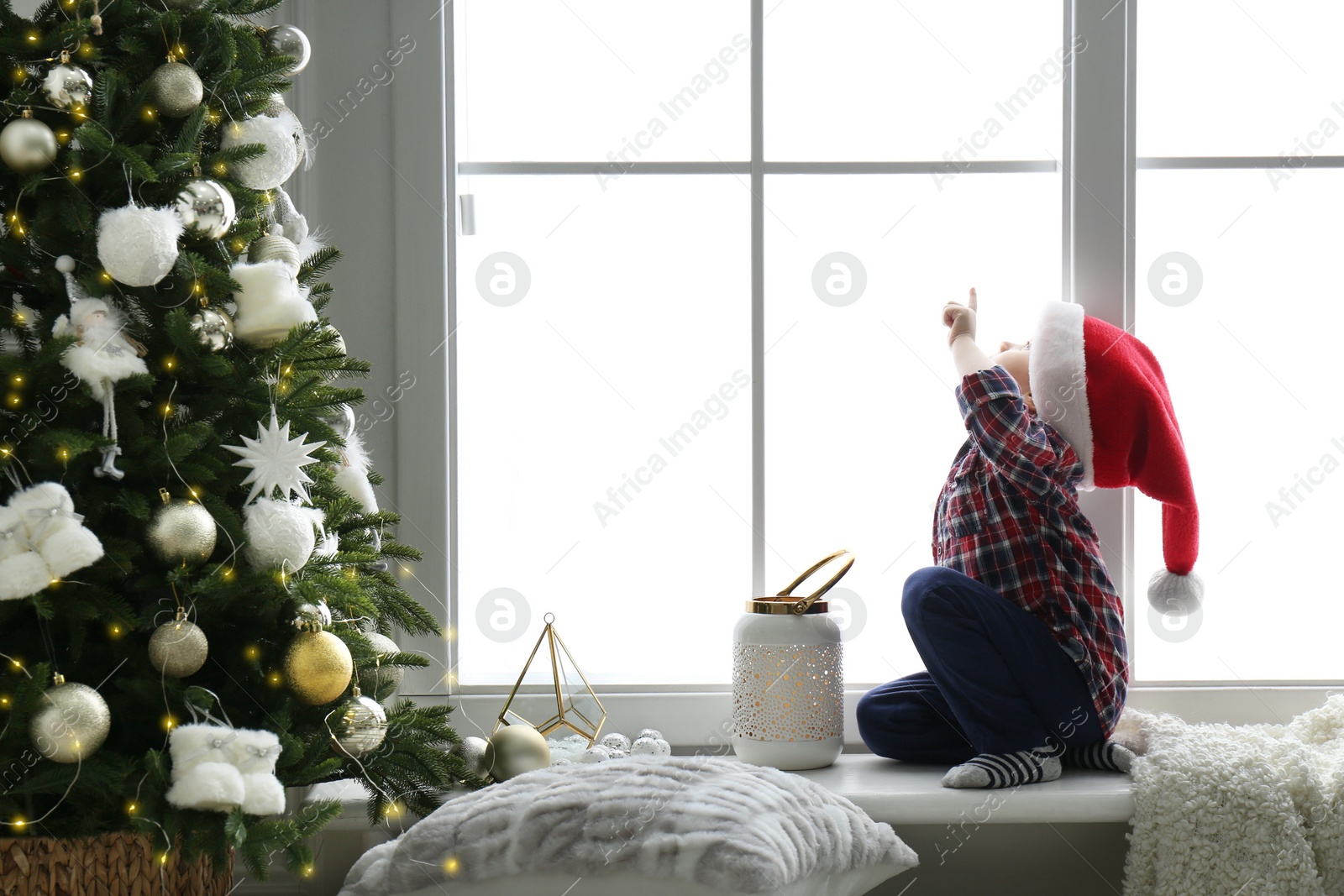 Photo of Cute little boy in Santa hat on window sill near Christmas tree at home