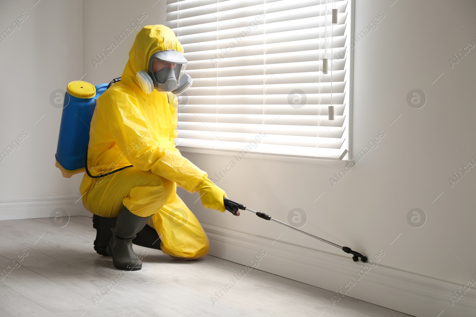 Photo of Pest control worker in protective suit spraying pesticide near window indoors