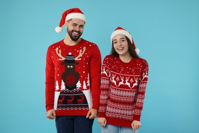 Photo of Happy young couple in Santa hats showing Christmas sweaters on light blue background