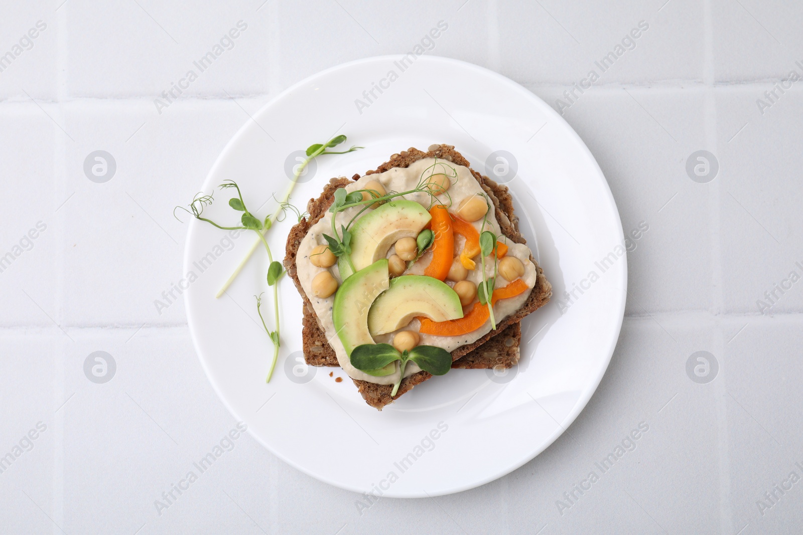 Photo of Tasty vegan sandwich with avocado, chickpeas and bell pepper on white tiled table, top view