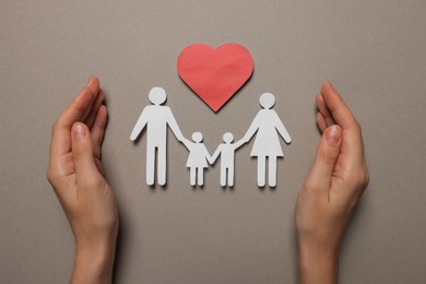 Photo of Woman protecting paper family figures and red heart on light grey background, top view. Insurance concept