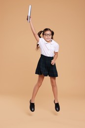 Photo of Cute schoolgirl holding book and jumping on beige background