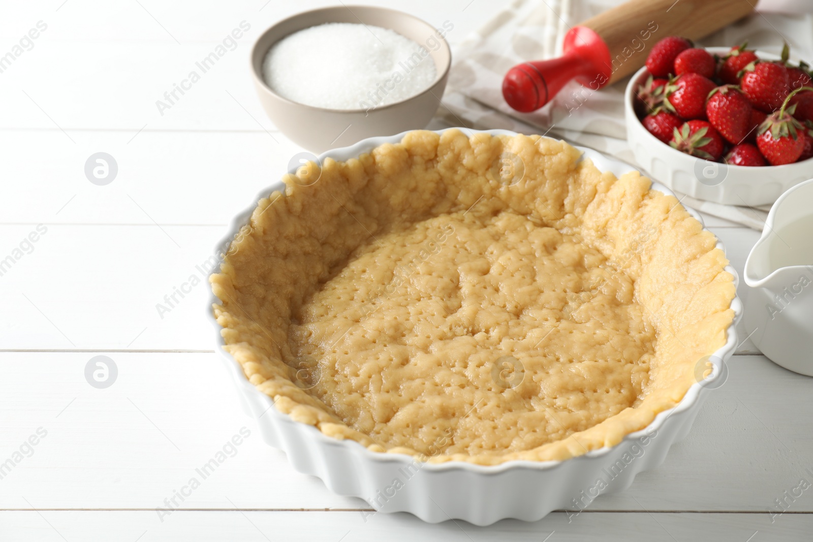 Photo of Making shortcrust pastry. Raw dough in baking dish, strawberries, sugar, rolling pin and milk on white wooden table