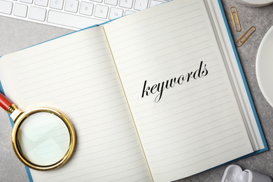 Image of Notebook with text KEYWORDS and magnifying glass on grey stone table, flat lay