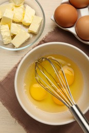Whisk and eggs in bowl, butter on wooden table, flat lay