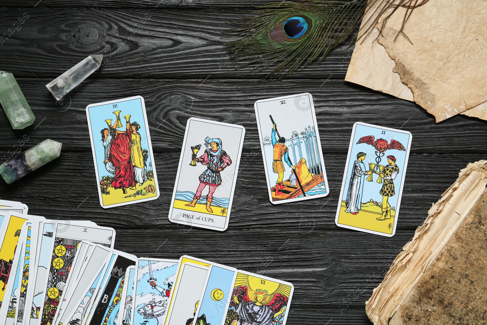 Photo of Tarot cards, old book, crystals and peacock feather on black wooden table, flat lay