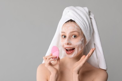 Emotional young woman washing face with brush and cleansing foam on grey background, space for text