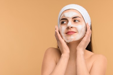 Young woman with headband washing her face on beige background, space for text