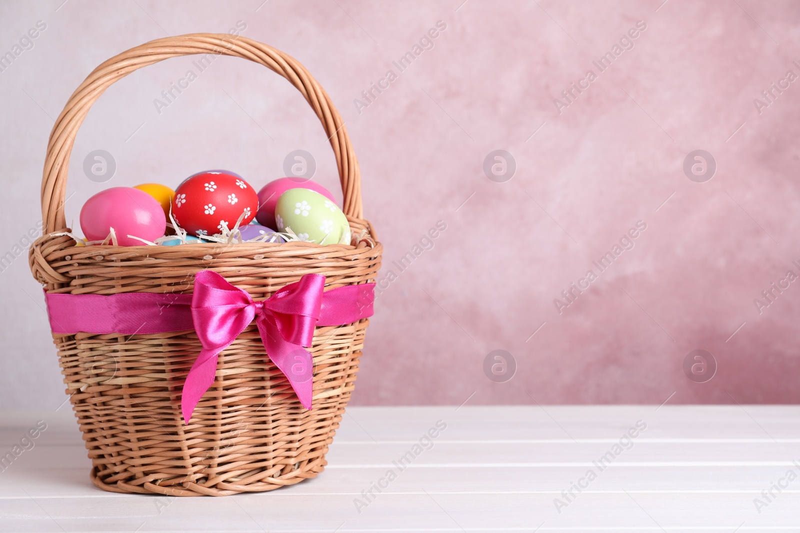 Photo of Wicker basket with bright painted Easter eggs on white wooden table against pink background. Space for text