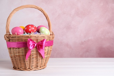 Wicker basket with bright painted Easter eggs on white wooden table against pink background. Space for text