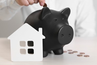 Photo of Man putting coin into piggy bank near house model at wooden table, closeup. Saving money concept