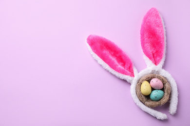 Photo of Headband with Easter bunny ears and dyed eggs in nest on violet background, flat lay. Space for text