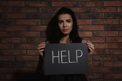 Abused young woman with sign HELP near brick wall. Domestic violence concept
